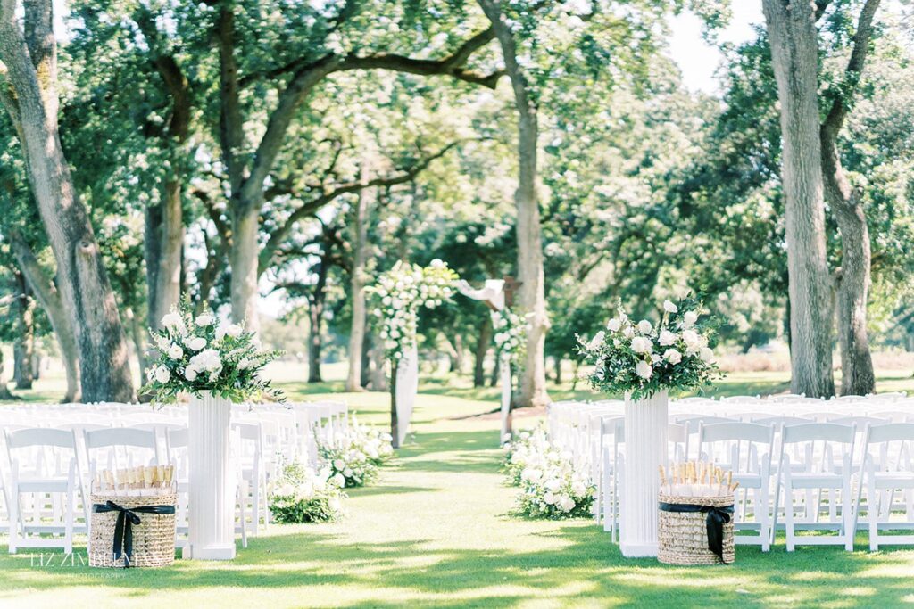 Outdoor wedding ceremony setup with white chairs, floral arrangements on pedestals, and a flower-adorned archway under tall trees.