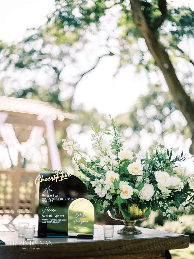 Outdoor wedding setup with a drink menu sign, floral arrangement of white roses, greenery, and candles on a wooden table. A pergola and trees are in the background.