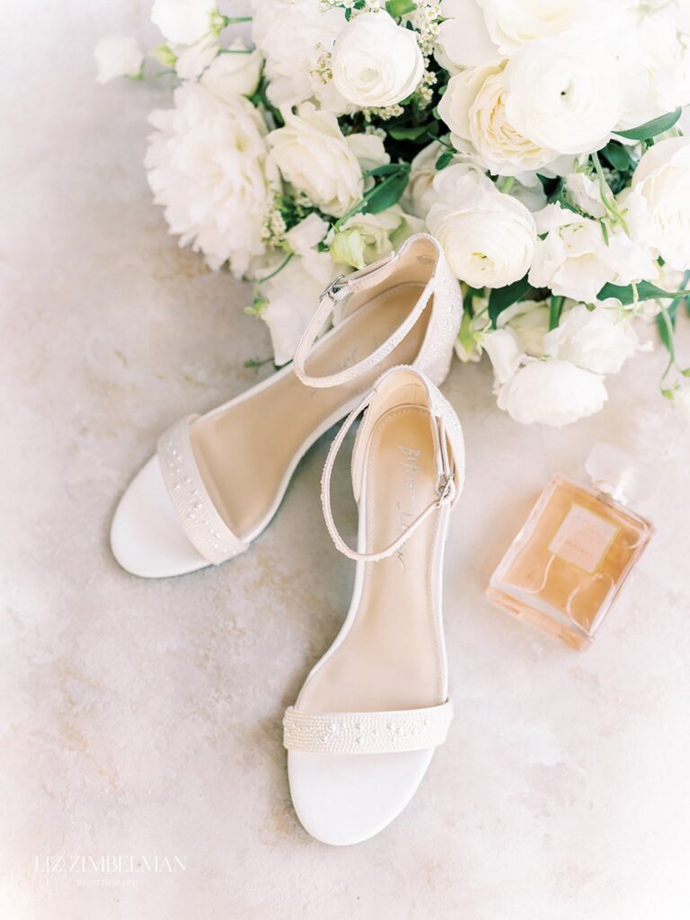 White open-toe bridal shoes with ankle straps on a textured surface, surrounded by white flowers and a bottle of perfume.