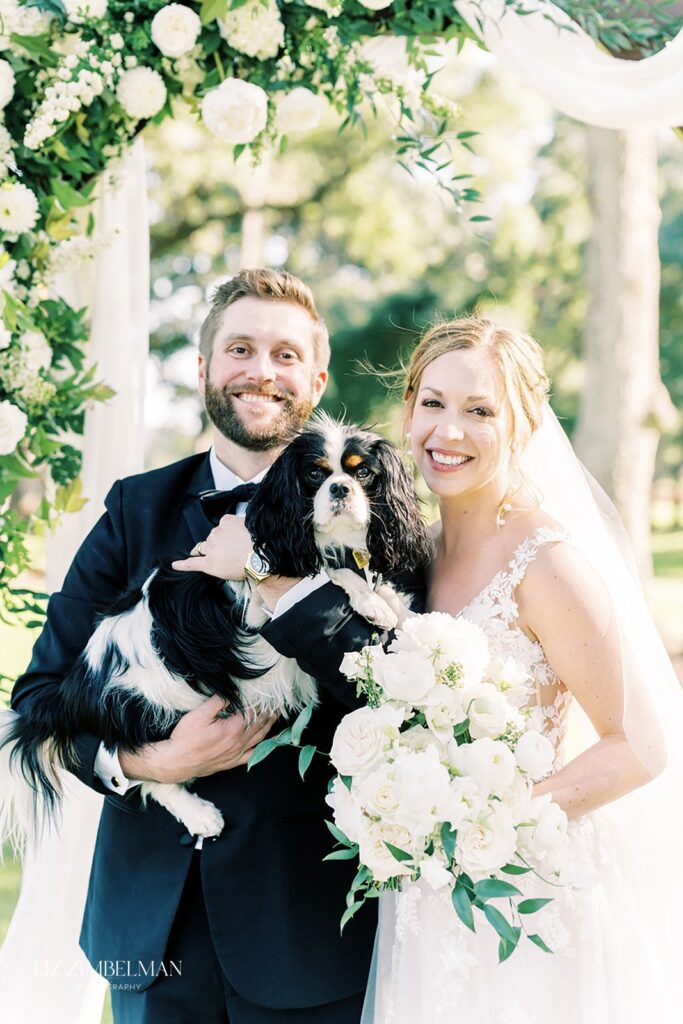 A couple in wedding attire poses outdoors, smiling and holding a black and white dog. Flowers and greenery embellish the scene.
