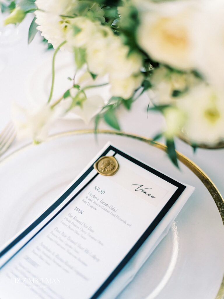 A close-up of a wedding menu on a plate. The menu features a gold wax seal and lists options for salad and main courses. White flowers are slightly blurred in the background.