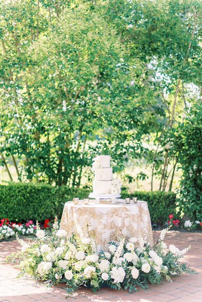 A tiered white cake on a decorated table with floral arrangements in an outdoor setting.