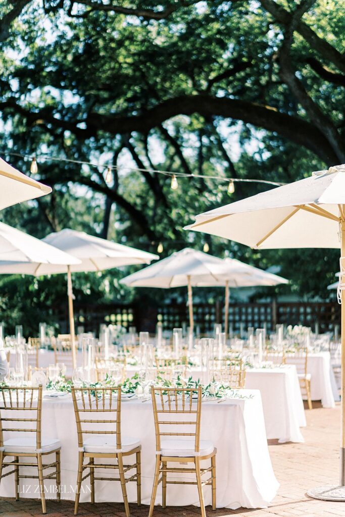 An outdoor event setup with white tablecloths, gold chairs, and white umbrellas under trees. String lights are visible above the tables.