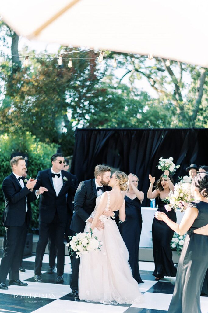 A couple shares a kiss at an outdoor wedding ceremony, surrounded by a cheering bridal party dressed in black formal attire.