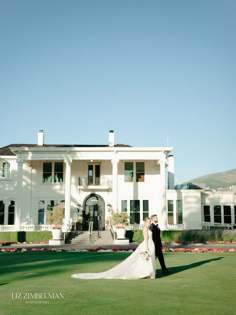 A bride and groom walk hand in hand on a manicured lawn in front of a large white mansion.
