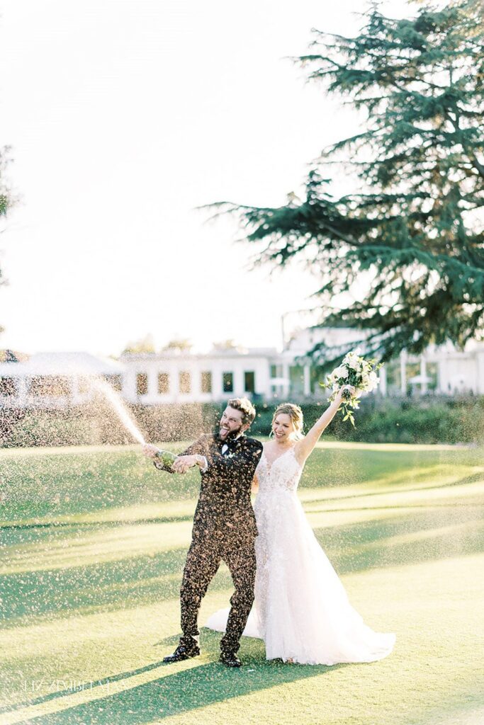 A couple in wedding attire celebrates outdoors by spraying champagne, with trees and a building in the background.