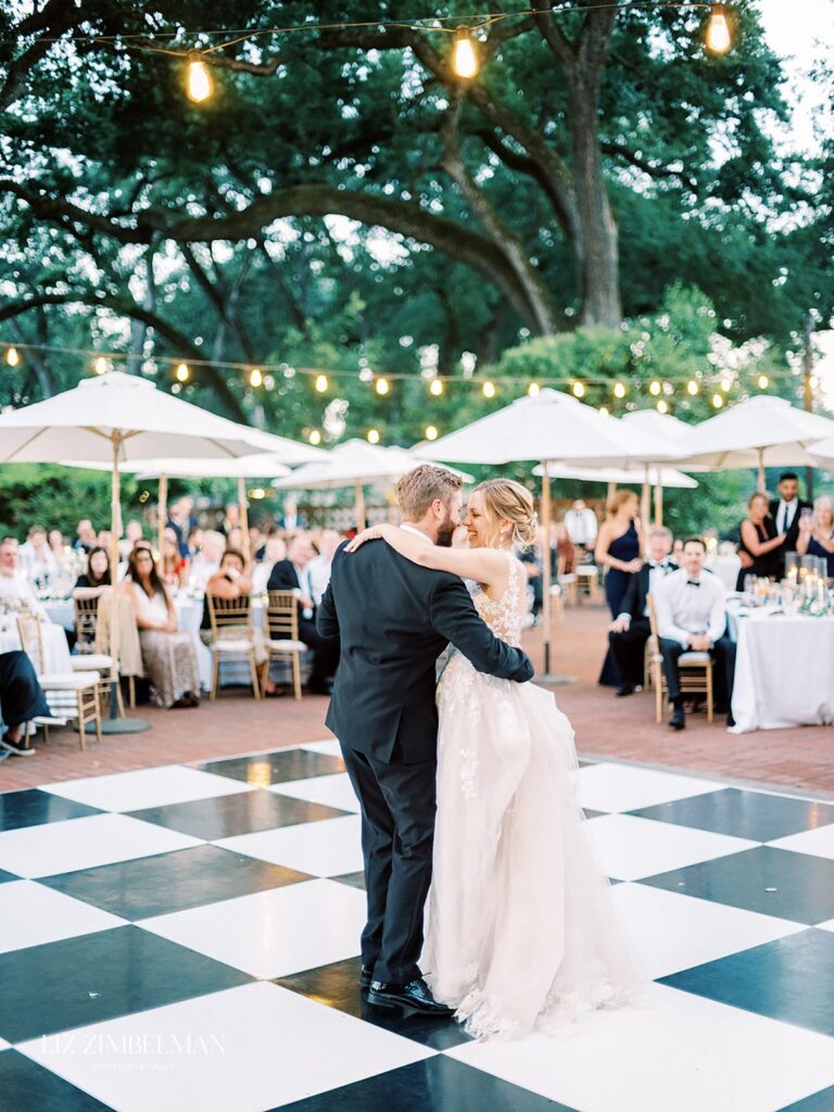 A couple dances on a checkered floor at an outdoor wedding reception with guests seated and string lights above.