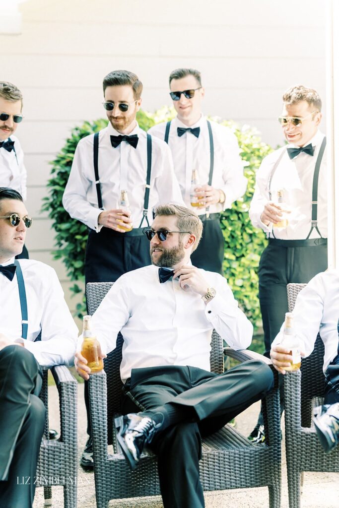 A group of men in sunglasses and formal attire, with bow ties and suspenders, sit and stand around outdoors, holding bottled drinks and engaging in conversation.