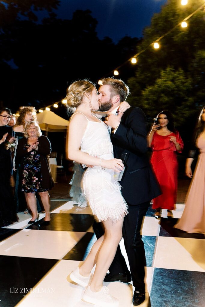 A couple dancing closely on a checkered floor under string lights, surrounded by people in formal attire.