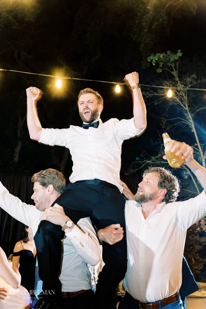 A man in a white shirt with his arms raised is lifted on the shoulders of two others at a nighttime outdoor event. One person holds a drink. String lights are visible in the background.