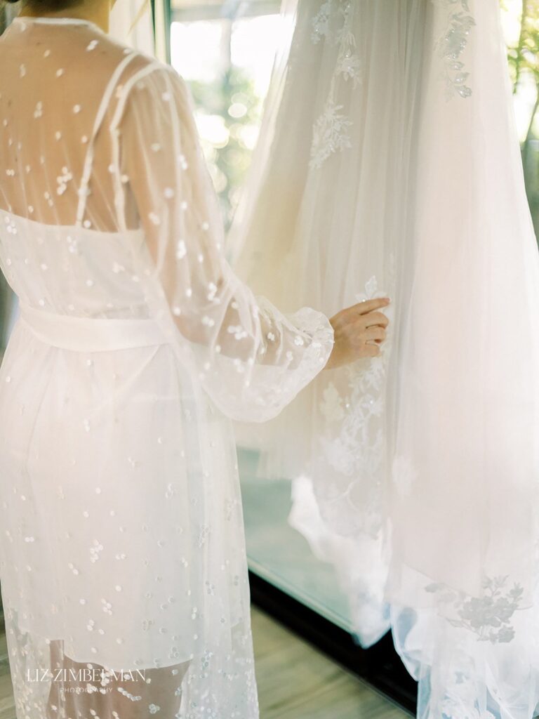 A person in a sheer white robe touches a hanging white lace dress in a softly lit room.
