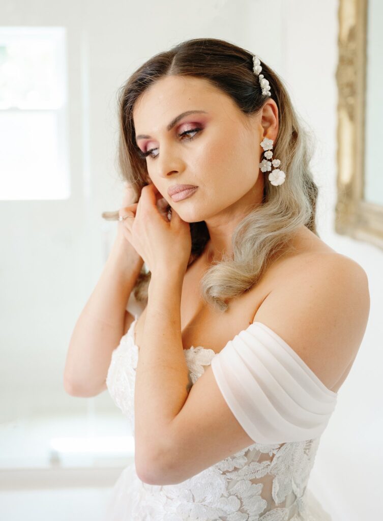 A bride with wavy hair, wearing a white off-shoulder gown and floral earrings, adjusts her hair in a bright room.