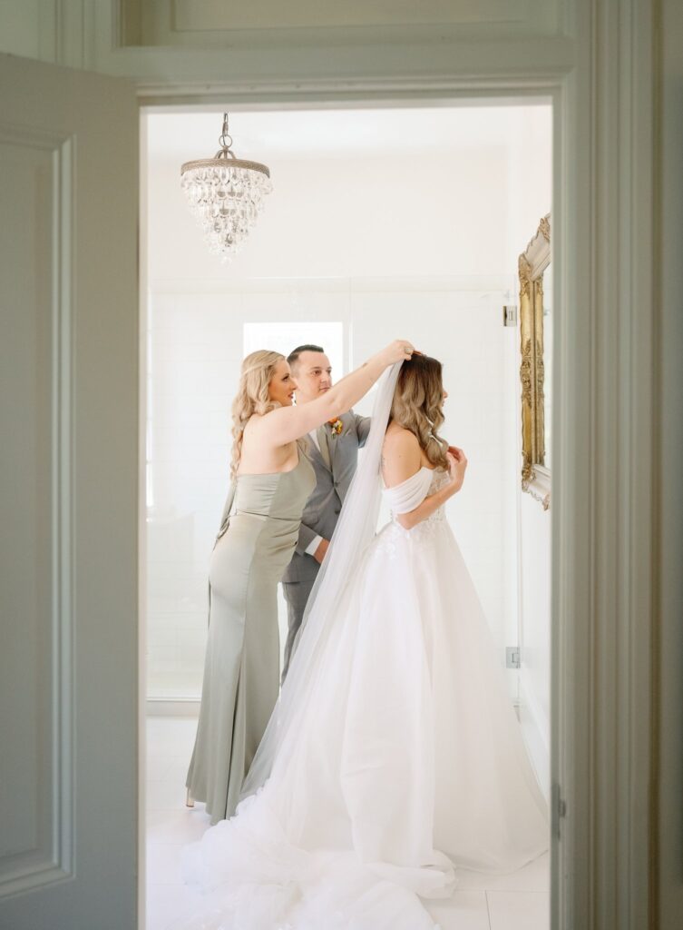 A woman adjusts another woman's veil while a man stands beside them in a sunlit room. They are all dressed formally, with the woman in white wearing a wedding dress.