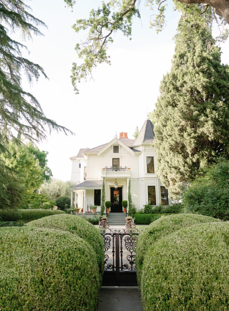 Victorian-style house with a pointed turret, surrounded by lush greenery and a wrought-iron gate in front.