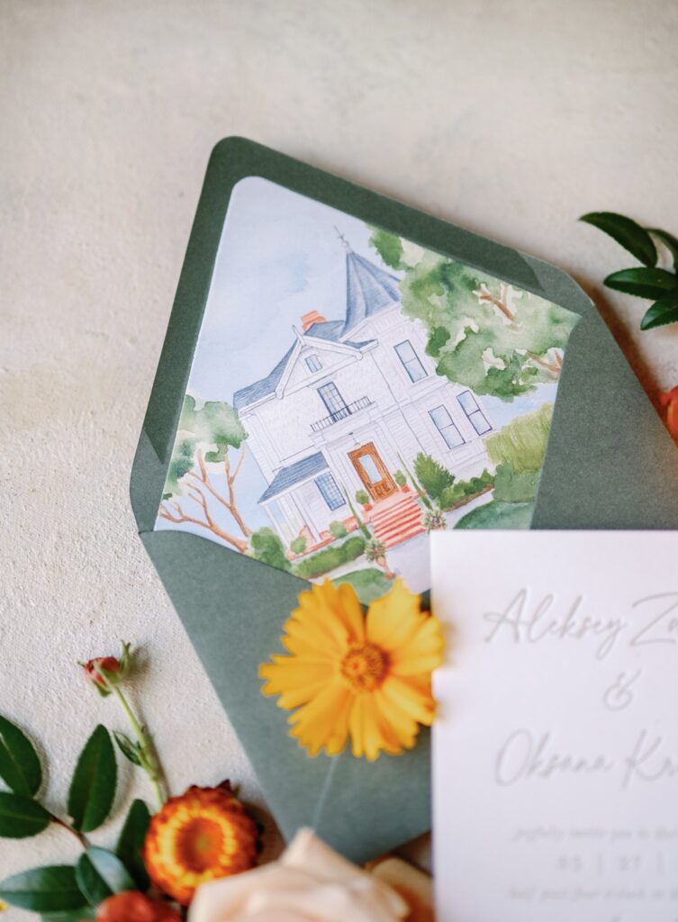 A green envelope with a watercolor illustration of a house is accompanied by a bouquet of orange flowers and a partially visible invitation card.