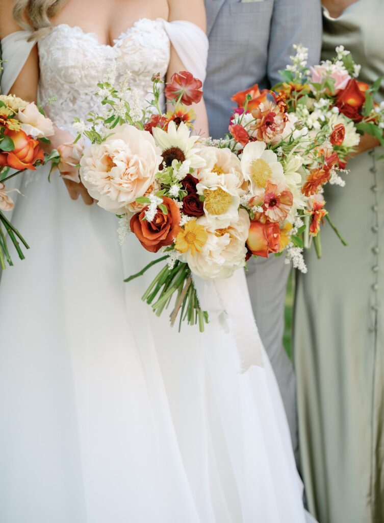 Three people holding bouquets of mixed flowers, including roses and peonies, with two in light-colored attire, one in a silver-gray suit.