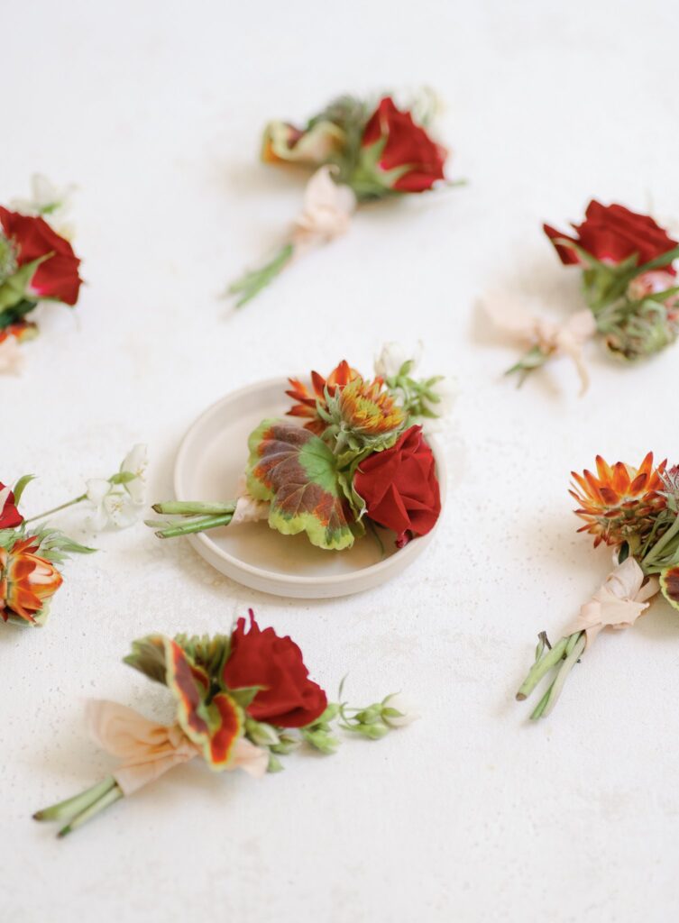 Several small floral arrangements with red roses and green leaves scattered on a white surface, with one placed on a small white plate in the center.