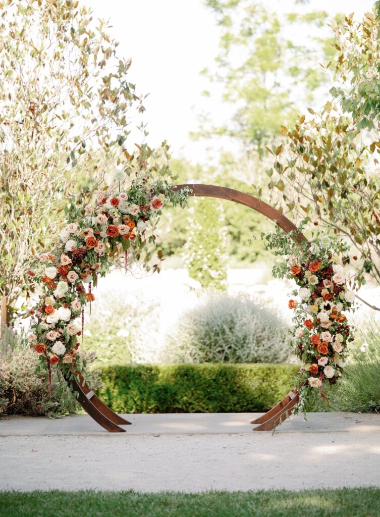 A circular wooden arch decorated with red, orange, and white flowers stands outdoors amid greenery.