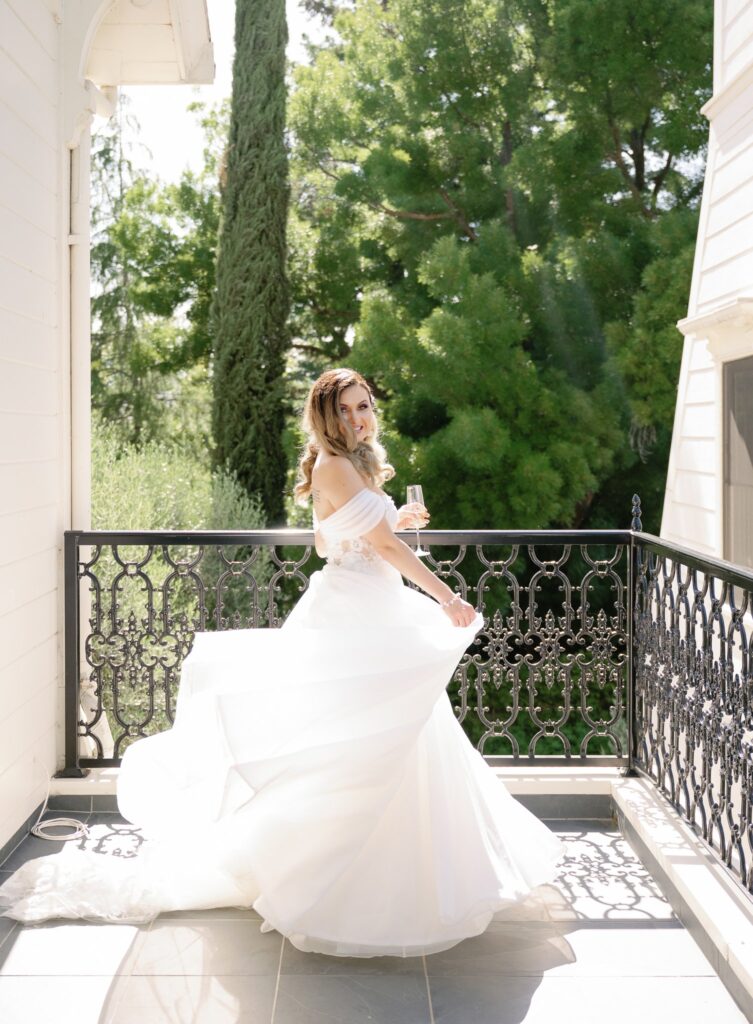 Bride in a white wedding dress holding a champagne glass stands on a sunlit balcony with trees in the background.