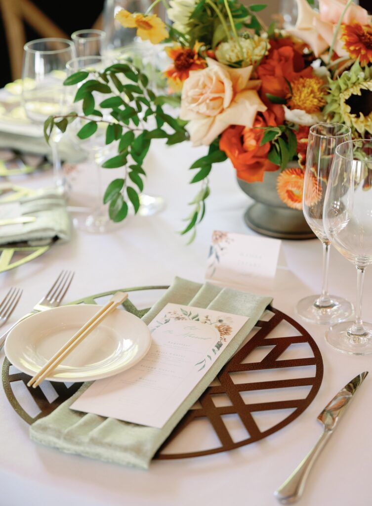 Elegant table setting with a floral centerpiece, a menu placed on a green napkin, a small plate with chopsticks, and glassware on a white tablecloth.