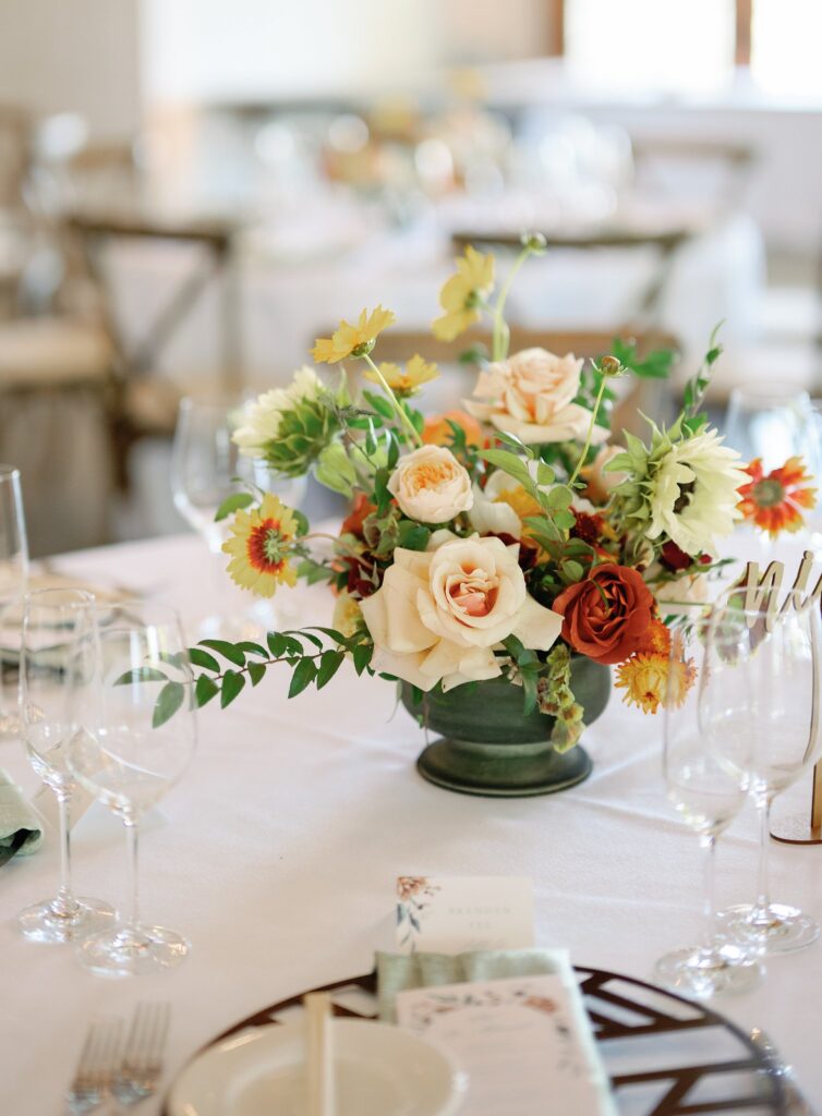 Table setting with floral centerpiece featuring cream and orange flowers, greenery, wine glasses, and a menu on a white tablecloth.