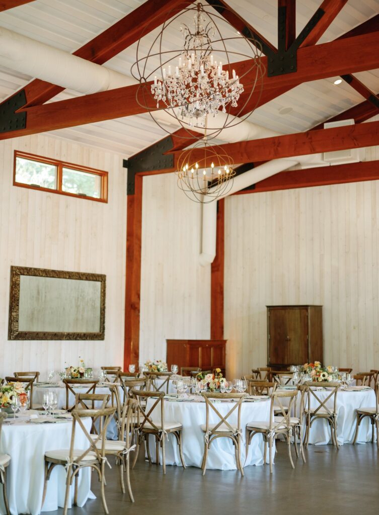 An elegant dining hall with wooden beams, round tables with white tablecloths, floral centerpieces, and a chandelier. Large mirror on the wall and a cabinet in the corner.