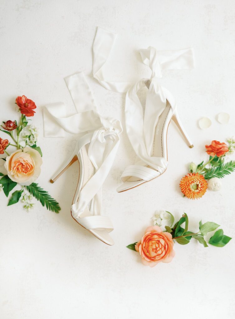 White high-heeled shoes with ribbon ties surrounded by orange and white flowers on a light background.