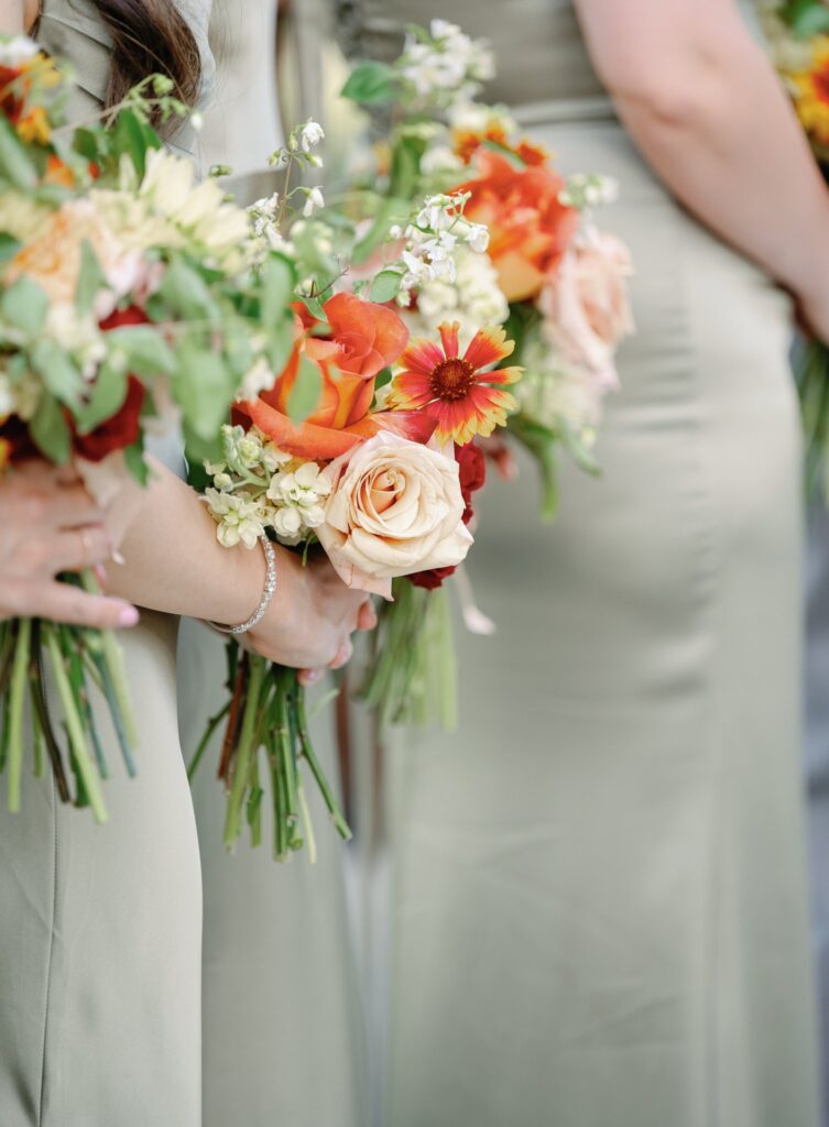 Bridesmaids in green dresses holding bouquets of orange, white, and yellow flowers.