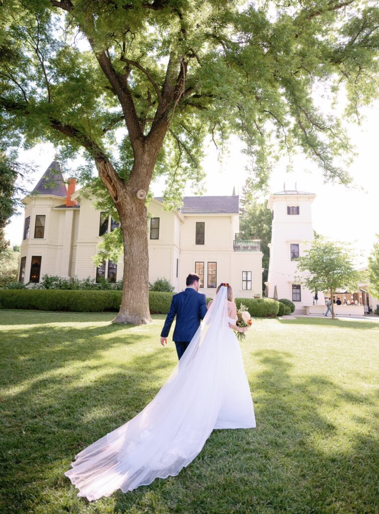 A bride and groom walk across a sunlit lawn towards a large, elegant house, with the bride's veil flowing behind them.