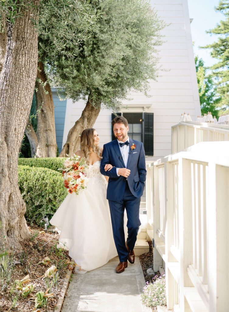 A bride and groom walk joyfully along a garden path. The bride holds a bouquet of flowers, and the groom wears a blue suit. Trees and a white building are in the background.