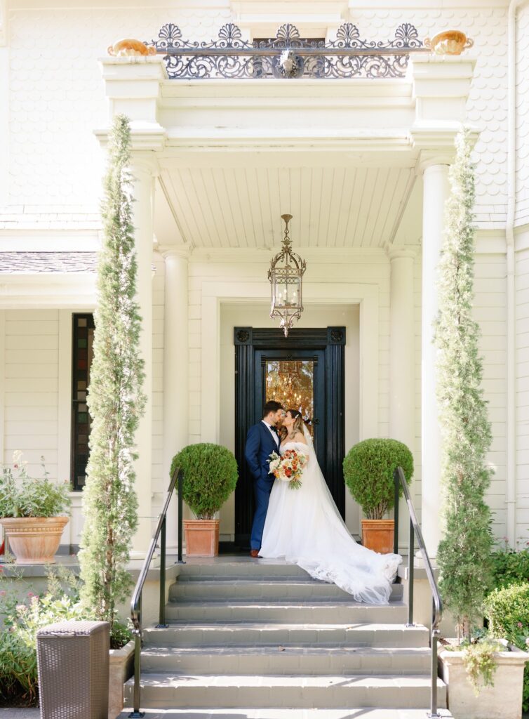 A bride and groom share a kiss on the steps of an elegant white building with black double doors, flanked by topiary plants and tall greenery.