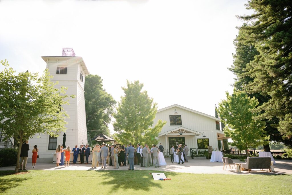 A group of people gathered outside a barn-style building with a tower on a sunny day. Trees and lawn surround the area.