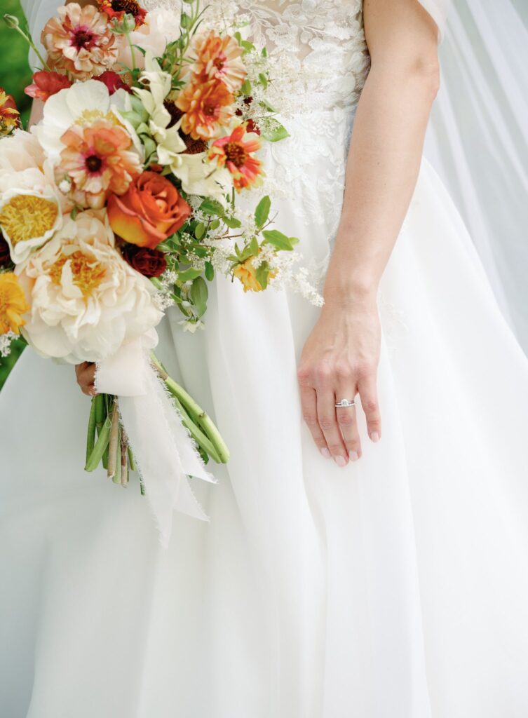 Bride in white dress holds a bouquet of orange, white, and yellow flowers. Her left hand shows a silver ring.