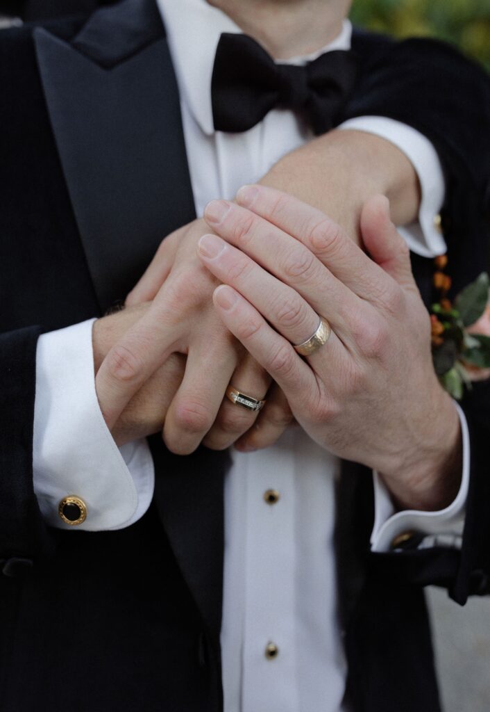 Two people wearing rings clasp hands. Both are in formal attire, including a suit jacket and dress shirt with cufflinks.