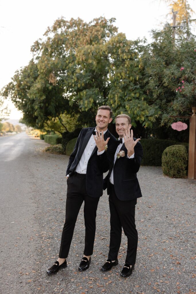 Two men in formal attire stand on a path, smiling and showing their rings. Trees and bushes line the path in the background.