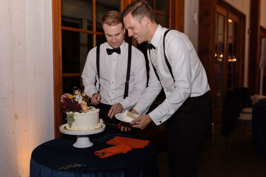 Two people in formal attire serve slices from a decorated cake on a stand over a dark blue tablecloth.