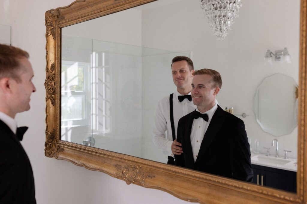 Two men in formal attire stand in front of a large ornate mirror. One adjusts the other's bow tie. A chandelier and a sink are visible in the background.