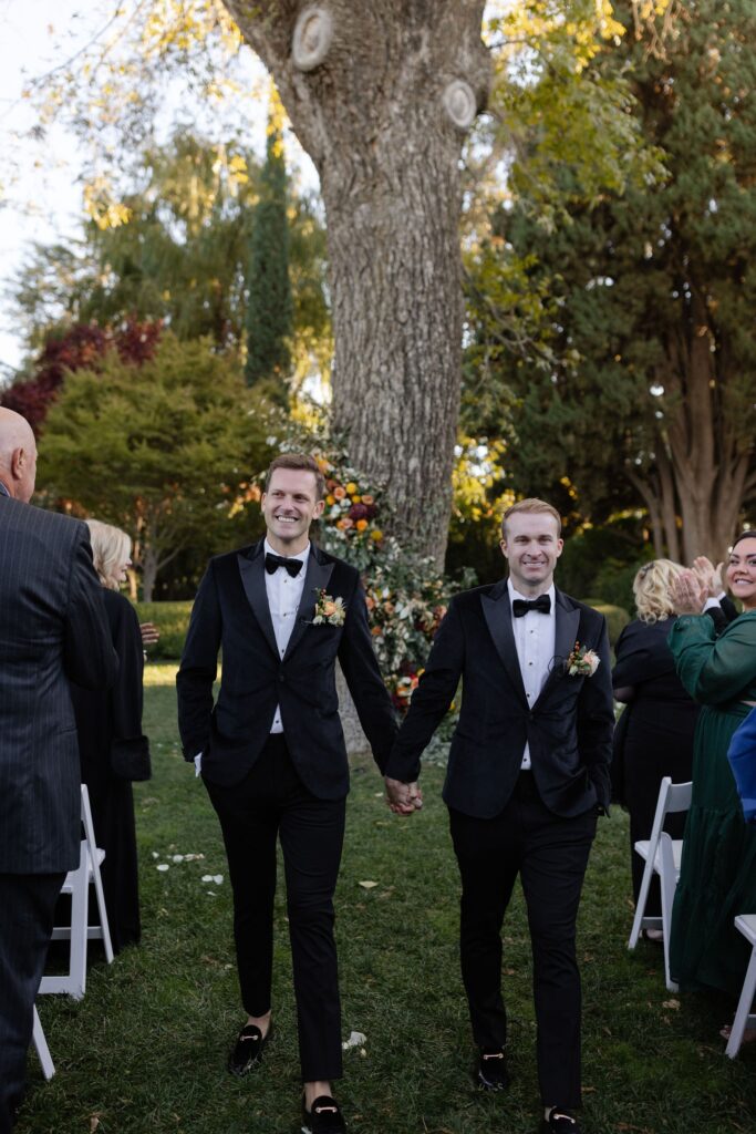 Two men in tuxedos hold hands and smile, walking outdoors in a garden ceremony with trees and seated guests around them.