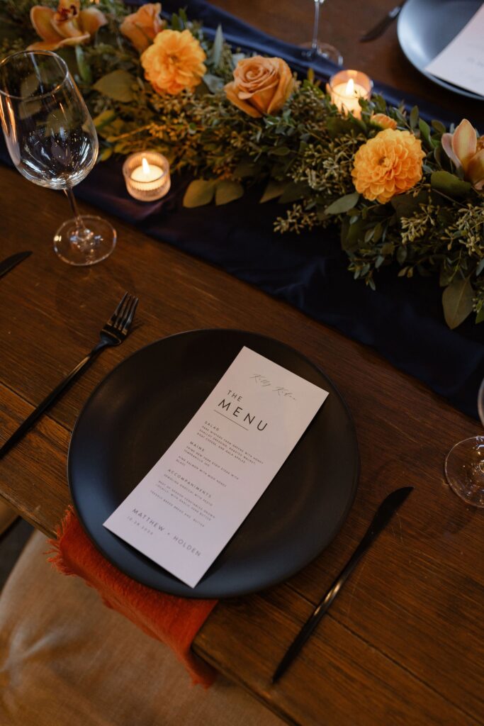 A black plate with a menu sits on a wooden table. Nearby are a black fork and knife, lit candles, and an arrangement of orange flowers and greenery.