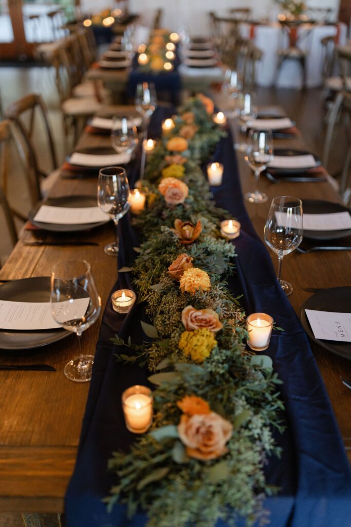 A wooden table is set for a formal meal with plates, glasses, and utensils. A centerpiece of candles, greenery, and orange flowers runs along a dark table runner.