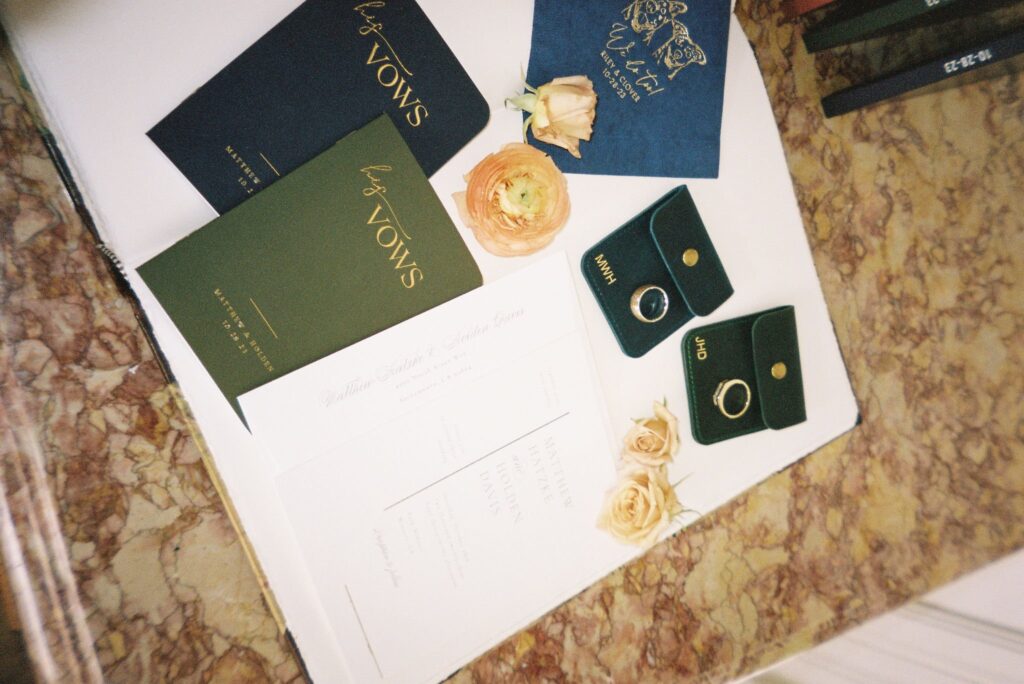 Wedding invitation, vow booklets, two rings in green cases, and beige roses on a marble surface.