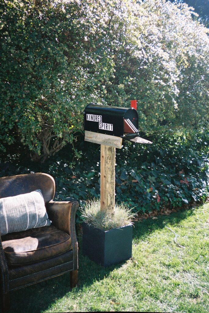 Mailbox labeled "Kindred Spirit" on a wooden post with an open lid, next to a brown armchair, set in a garden with greenery.