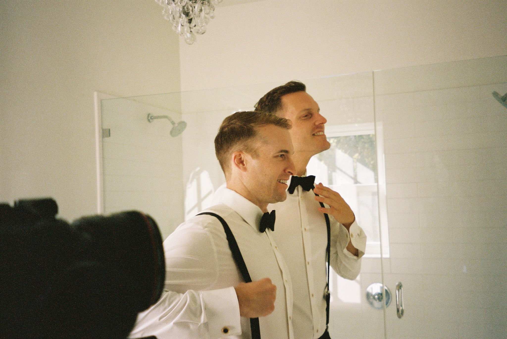 Two men in white dress shirts and bow ties stand in front of a mirror adjusting their outfits, smiling. They appear to be getting ready for a formal event.