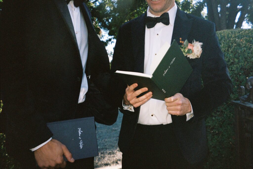 Two men in suits holding green booklets, standing outdoors. One man has a floral boutonniere.