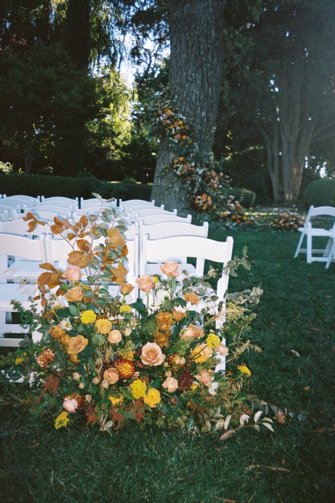 White folding chairs arranged outdoors with lush floral decorations of orange and yellow flowers on a grassy area, surrounded by tall trees.