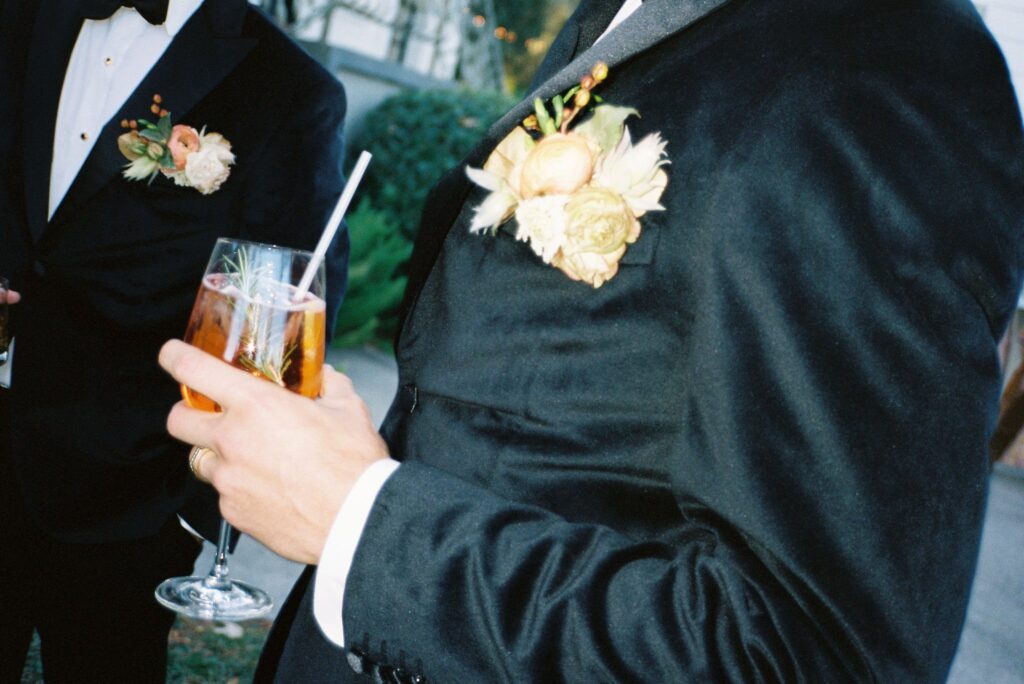 Two people in formal black attire hold drinks, each with a floral boutonniere. One person is holding a glass with a straw.