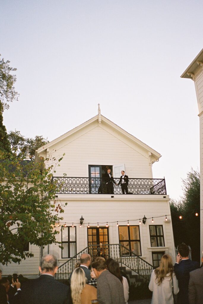 A crowd of people stands in front of a two-story house with a balcony, where two people are speaking to the gathered audience.