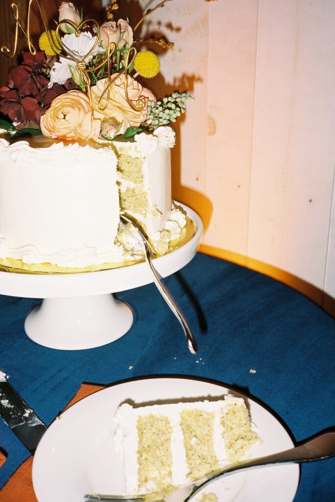 A sliced cake with white icing and floral decoration sits on a stand, next to a plate with a piece of the cake, on a blue tablecloth.