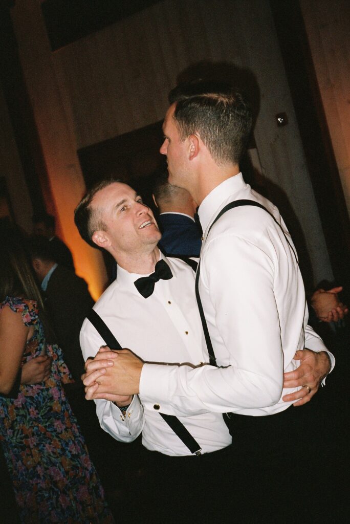 Two people in formal attire dance together, both wearing white shirts and dark suspenders. They are surrounded by other people in a dimly lit room.