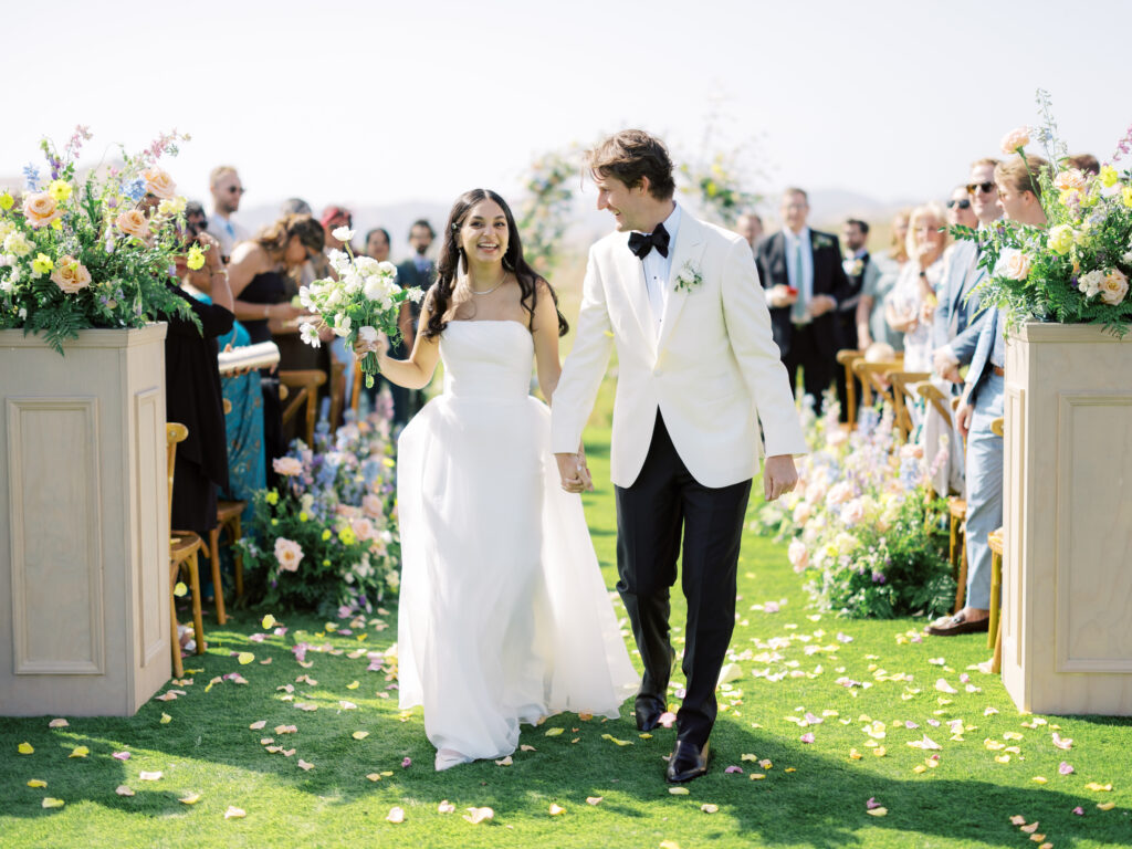A bride and groom walk down an outdoor aisle, surrounded by guests and floral arrangements.